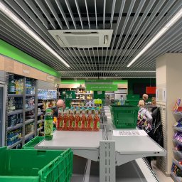 Mistre Group sp. z o.o. - Nowoczesny sufit rastrowy z oświetleniem liniowym w supermarkecie, klimatyzacja centralna, półki z napojami i przekąskami. Widoczne osoby robiące zakupy.