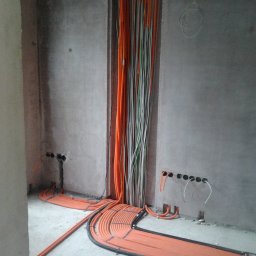 Mistre Group sp. z o.o. - Pomarańczowe kable elektryczne ułożone wzdłuż ściany w surowym wnętrzu. Widoczne otwory w ścianie i pionowe wiązki kabli. Instalacja elektryczna w trakcie realizacji.