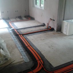 Mistre Group sp. z o.o. - Pomarańczowe i czarne kable elektryczne ułożone na podłodze w trakcie instalacji, widoczne puszki podtynkowe i materiały izolacyjne. Etap prac przed wylaniem posadzki.