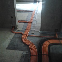 Mistre Group sp. z o.o. - Pomarańczowe kable elektryczne równolegle ułożone na podłodze w surowym wnętrzu, zabezpieczone czarnymi opaskami, prowadzone między betonowymi filarami.