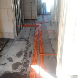 Mistre Group sp. z o.o. - Pomarańczowe kable elektryczne ułożone na podłodze w surowym wnętrzu, widoczne profile ścian działowych i postać w oddali, oświetlone jasnym światłem.