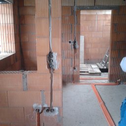 Mistre Group sp. z o.o. - Surowy stan wnętrza z ceglanymi ścianami, widoczne przewody elektryczne i rury instalacyjne w podłodze. Etap prac przed wykończeniem.