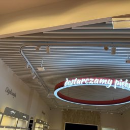 Mistre Group sp. z o.o. - Sufit rastrowy z oświetleniem punktowym i neonowym napisem 'dostarczamy piękno' w jasnym wnętrzu sklepu. Widoczny system kamer i elementy dekoracyjne.
