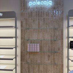 Mistre Group sp. z o.o. - Beżowa ściana z płytek, podświetlony napis 'Nasi eksperci polecają', szklane półki z produktami. Widok na wykończone wnętrze salonu.