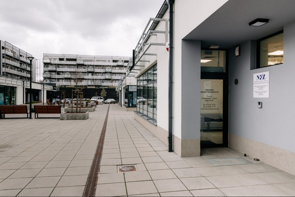 Elewacja budynku usługowego z oknami i drzwiami, widok z zewnątrz. Szara kolorystyka, nowoczesna architektura, chodnik z kostki brukowej. Tabliczka informacyjna NFZ.