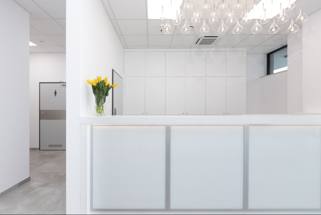 Białe wnętrze biura z recepcją, żółte tulipany w wazonie, minimalistyczny design, oświetlenie sufitowe, drzwi z oznaczeniem toalety w tle.