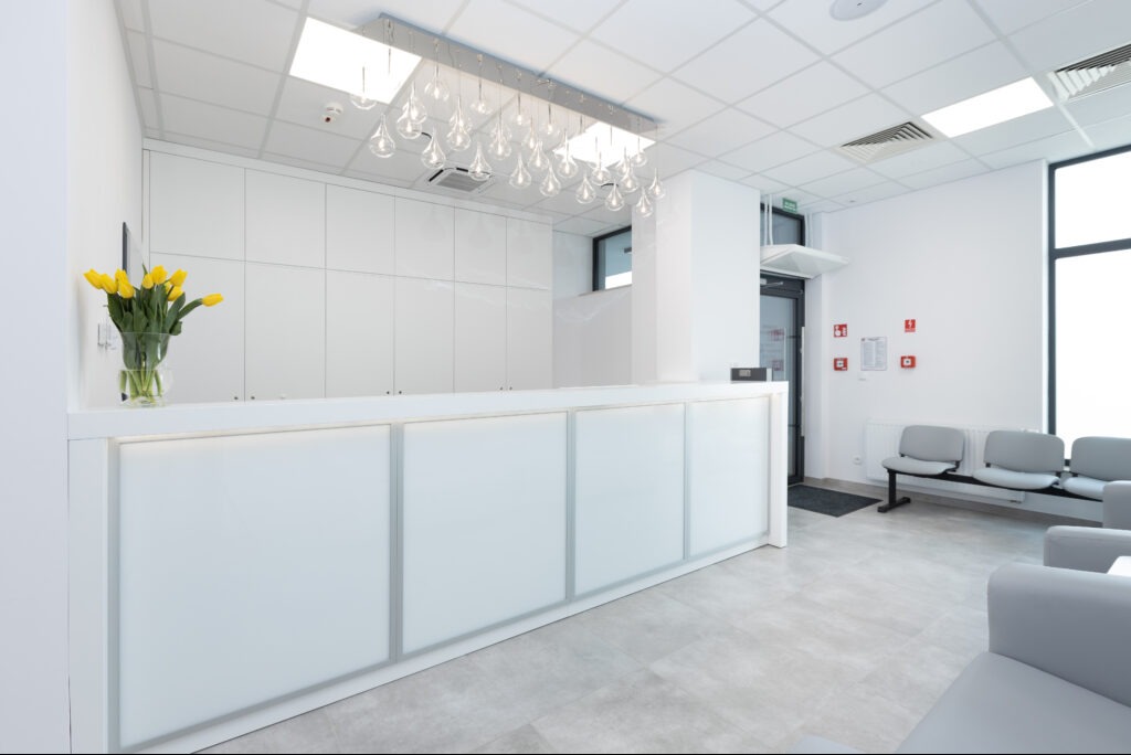 Nowoczesne wnętrze biura z recepcją i strefą oczekiwania. Minimalistyczny design, białe meble, szare podłogi, żółte tulipany w wazonie. Oświetlenie sufitowe.