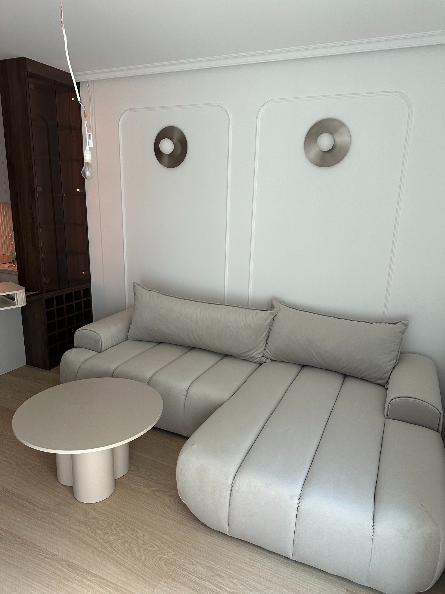 Stylowe wnętrze: jasnoszara sofa z okrągłym stolikiem na drewnianej podłodze. Dekoracyjne panele ścienne z oświetleniem i wbudowany regał z winami.