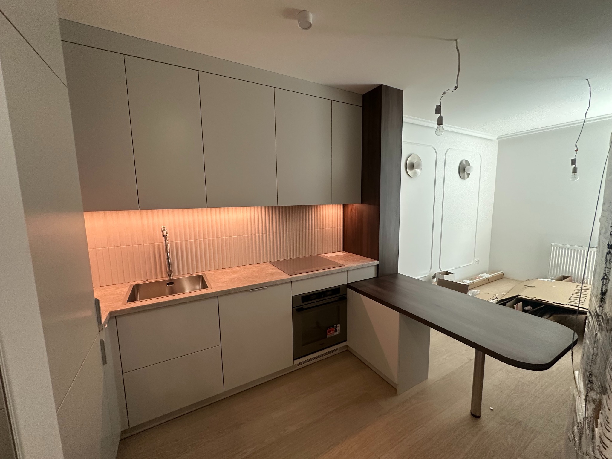 Nowoczesna kuchnia z szarymi szafkami, podświetlanym blatem i ciemnym drewnianym barkiem. Widoczne niedokończone wykończenia w tle.