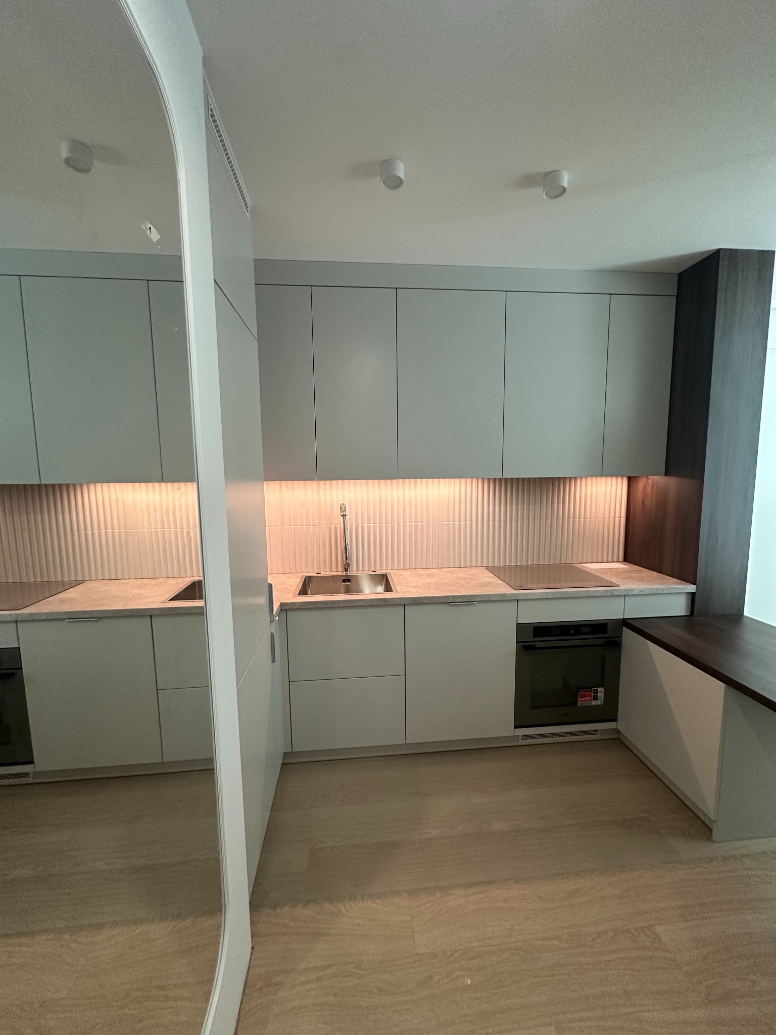 Nowoczesna kuchnia w odcieniach szarości z podświetlanymi płytkami ściennymi i drewnianą podłogą. Element lustra po lewej stronie kadru.