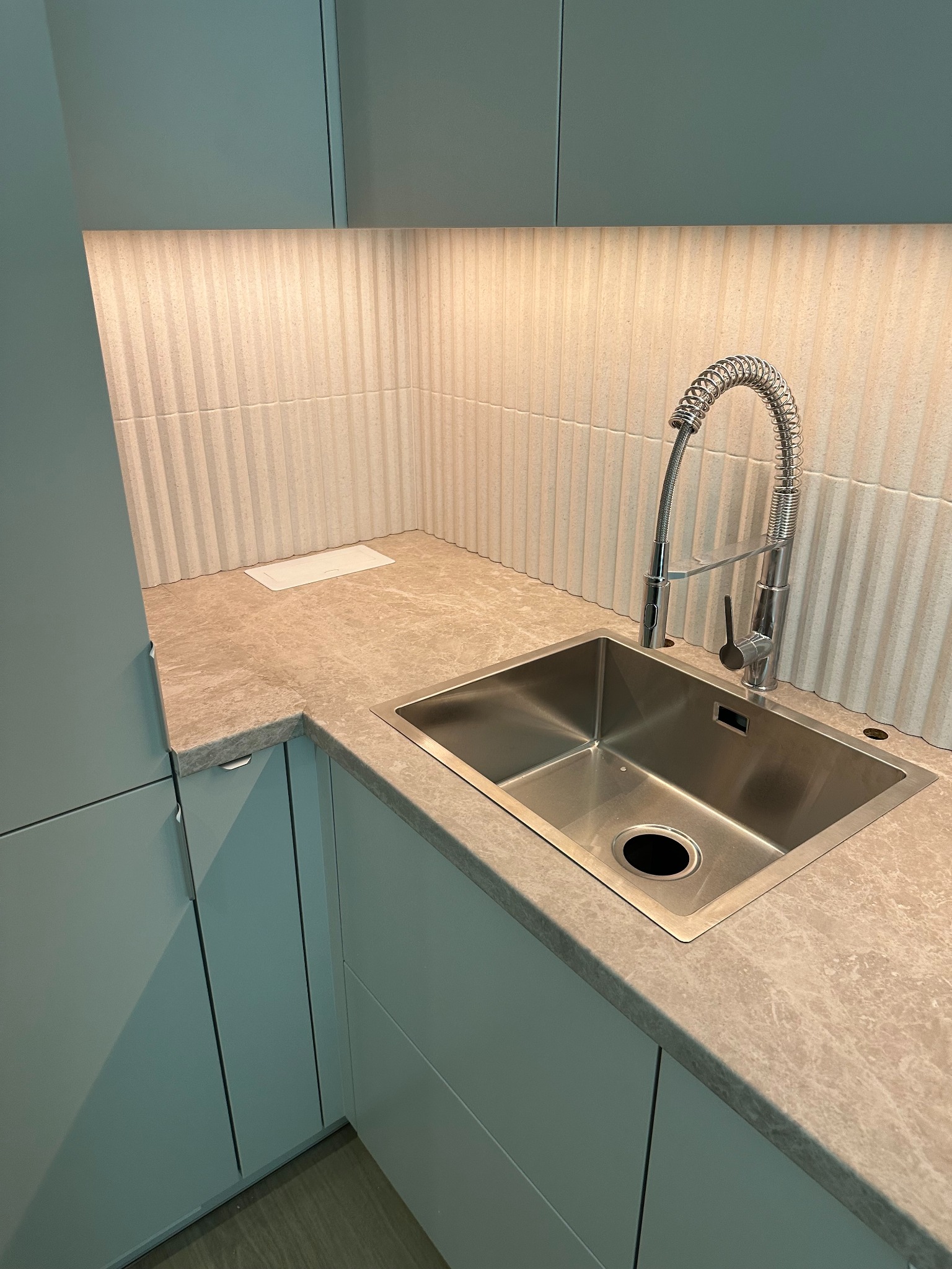 Nowoczesna kuchnia z szarymi szafkami, marmurowym blatem i stalowym zlewem z baterią sprężynową. Oświetlenie LED podkreśla pionowe płytki na ścianie.