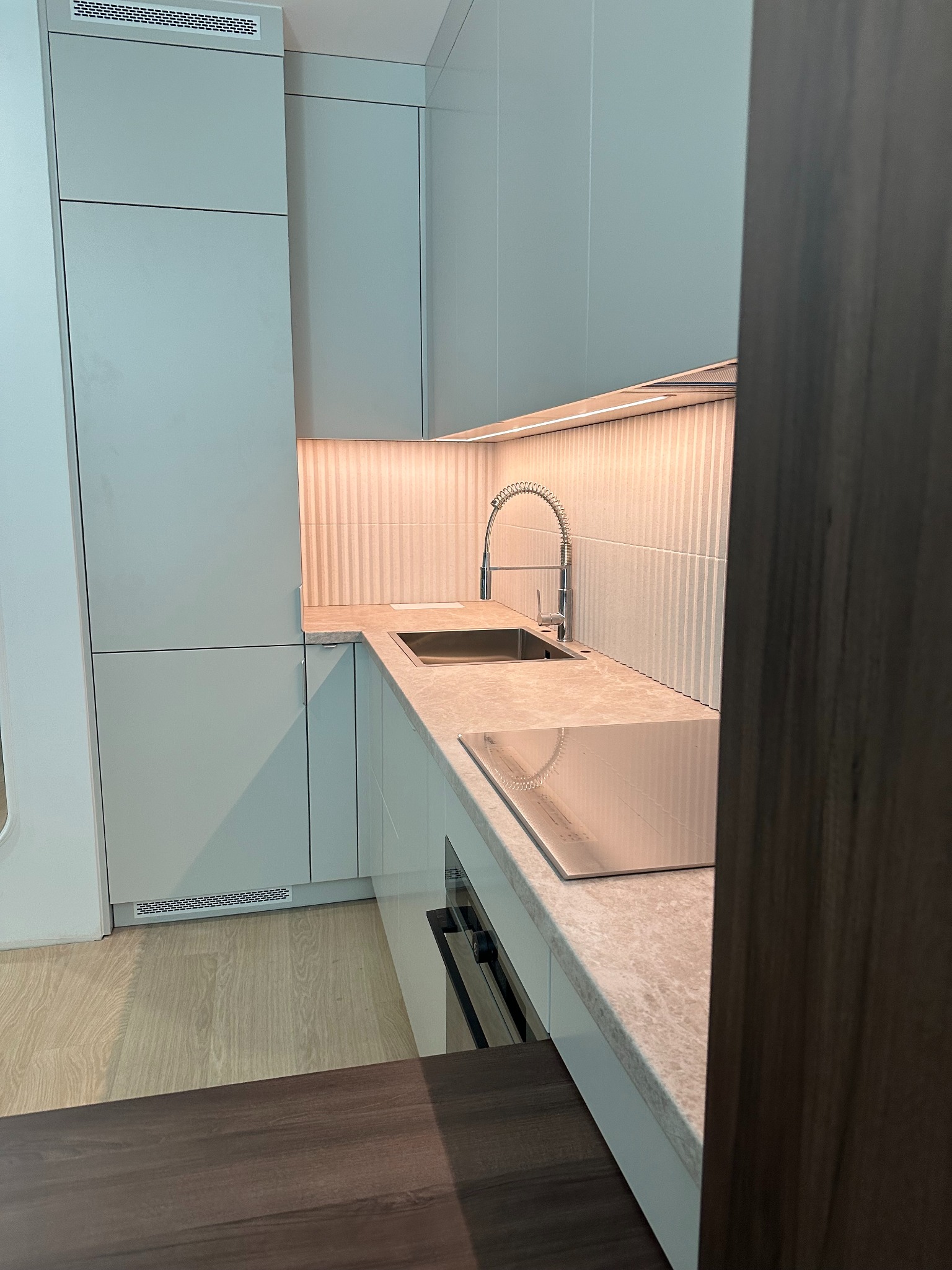 Nowoczesna, minimalistyczna kuchnia z jasnymi szafkami, marmurowym blatem i oświetleniem LED, widziana z niskiego kąta, z fragmentem ciemnego blatu w tle.