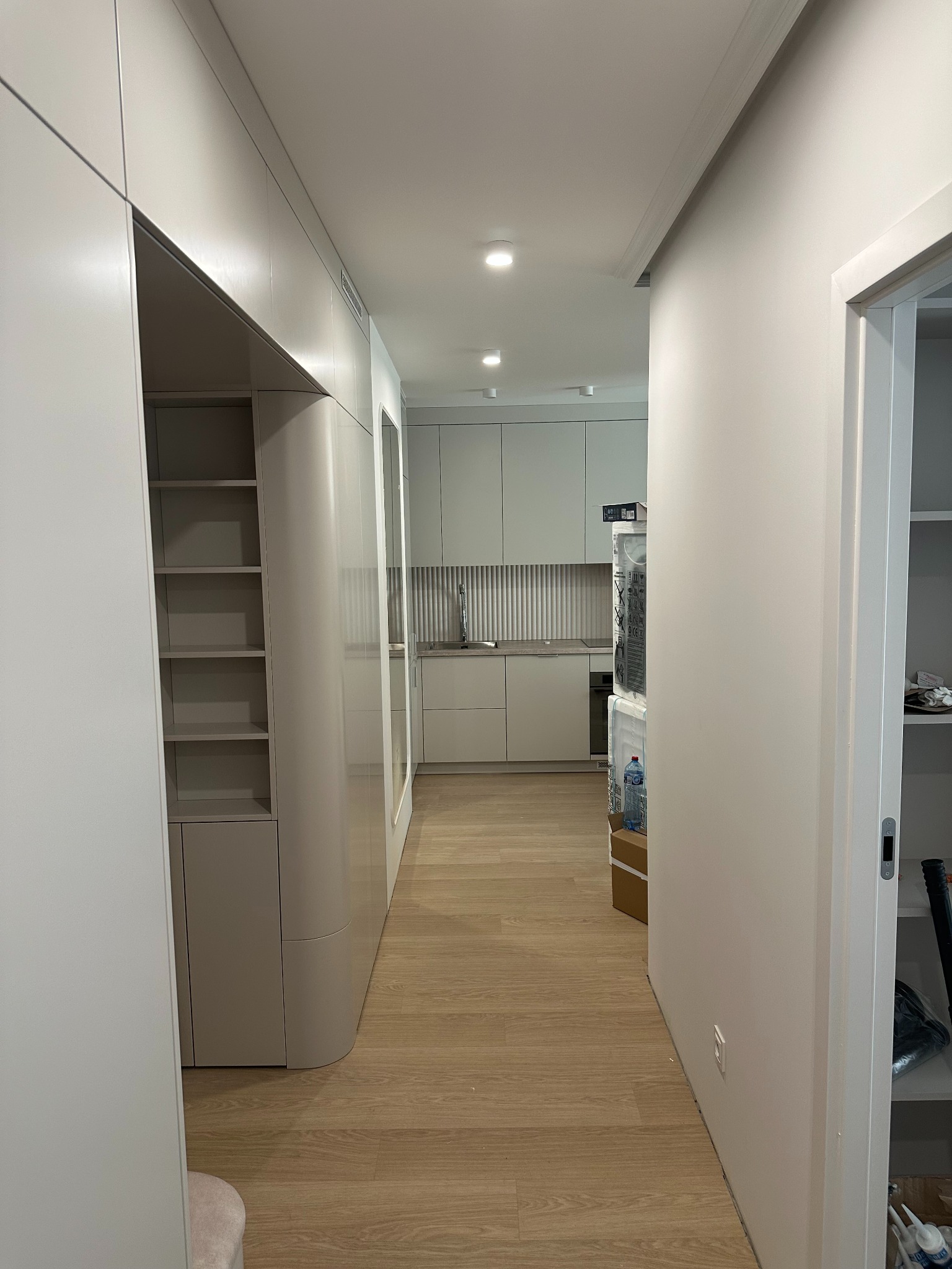 Nowoczesne wykończenie wnętrza: korytarz z zabudową meblową w odcieniach szarości, drewniana podłoga i widok na kuchnię z ryflowanym panelem ściennym.