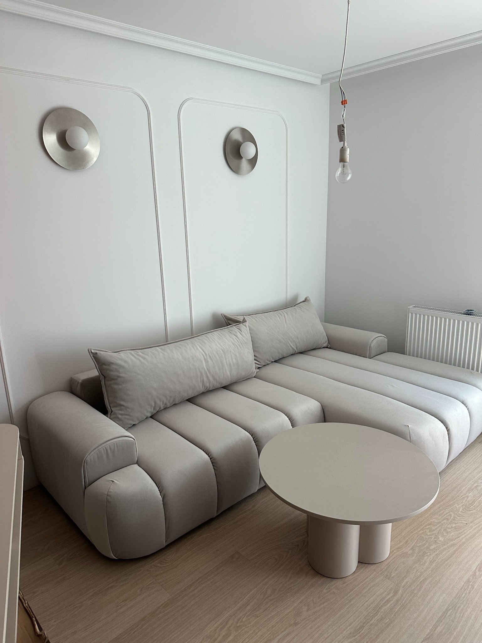 Stylowe wnętrze z beżową sofą modułową i okrągłym stolikiem. Na ścianie ozdobne panele z okrągłymi lampami. Widoczna niedokończona instalacja elektryczna.