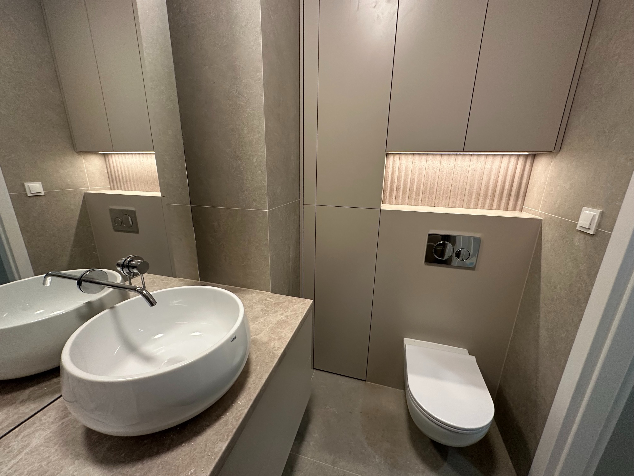 Nowoczesna łazienka w beżach: owalna umywalka Cielo, podświetlane szafki, płytki imitujące kamień. Minimalistyczny design, eleganckie wykończenie.