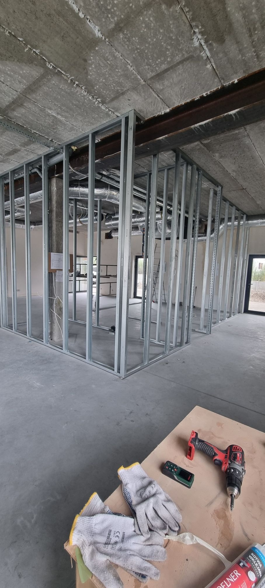 Montaż szkieletu ścian działowych z profili stalowych w surowym wnętrzu. Na pierwszym planie narzędzia: wkrętarka, dalmierz i rękawice robocze.