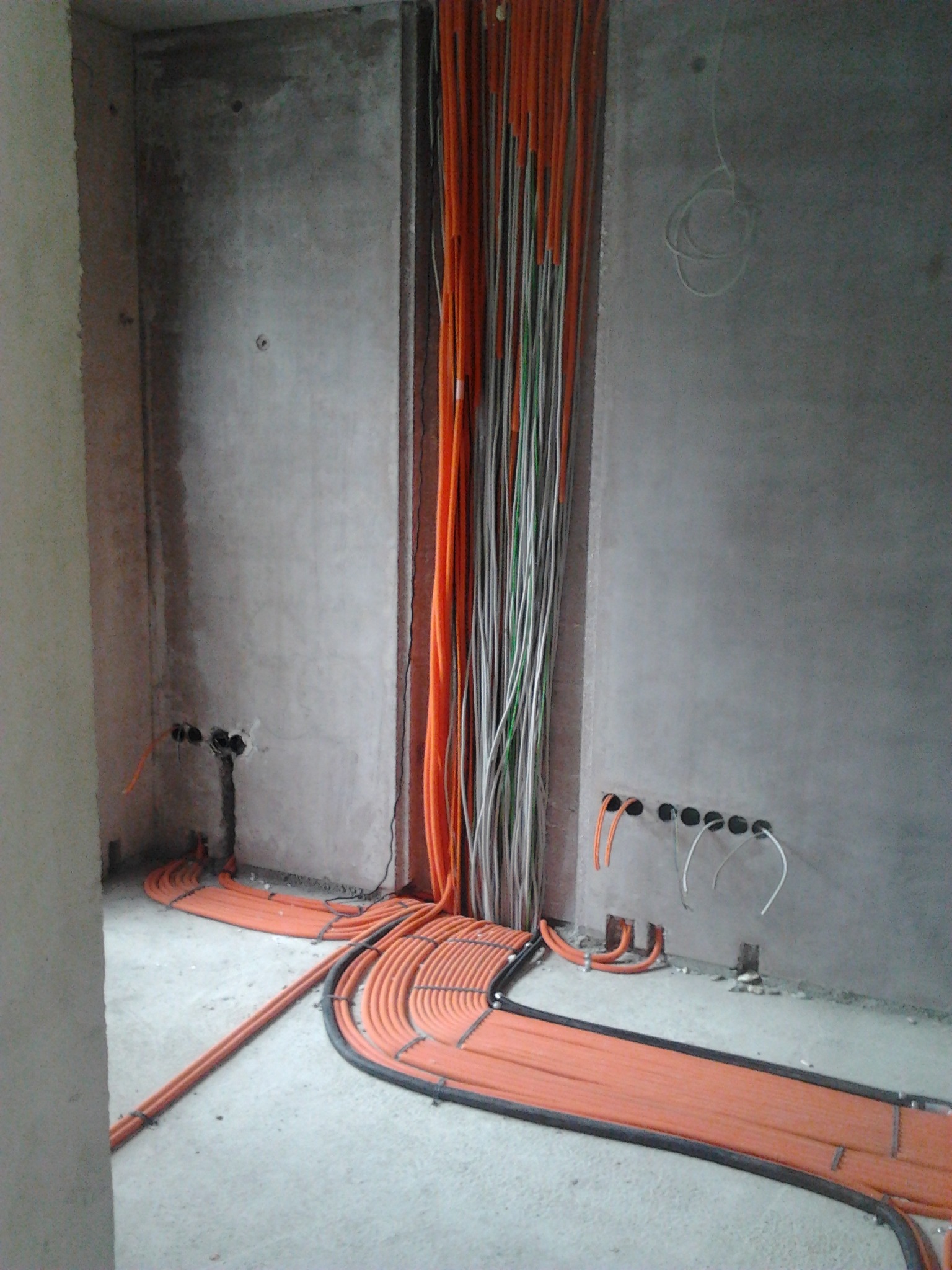 Pomarańczowe kable elektryczne ułożone wzdłuż ściany w surowym wnętrzu. Widoczne otwory w ścianie i pionowe wiązki kabli. Instalacja elektryczna w trakcie realizacji.
