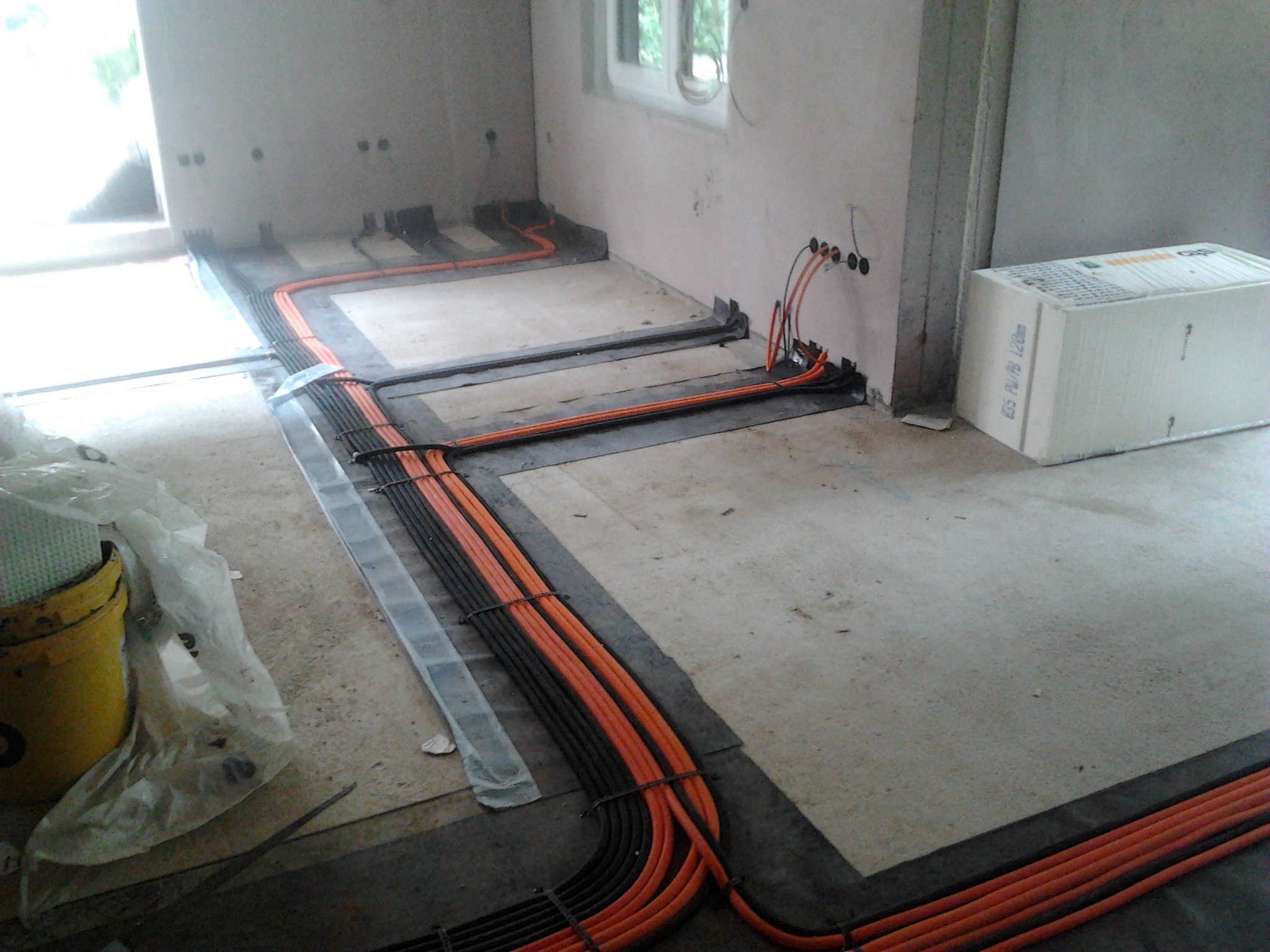 Pomarańczowe i czarne kable elektryczne ułożone na podłodze w trakcie instalacji, widoczne puszki podtynkowe i materiały izolacyjne. Etap prac przed wylaniem posadzki.
