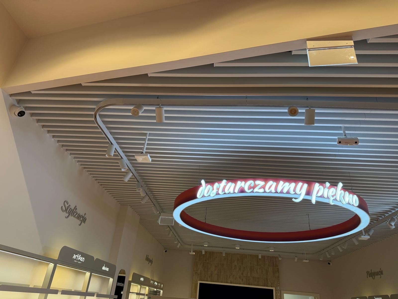 Sufit rastrowy z oświetleniem punktowym i neonowym napisem 'dostarczamy piękno' w jasnym wnętrzu sklepu. Widoczny system kamer i elementy dekoracyjne.