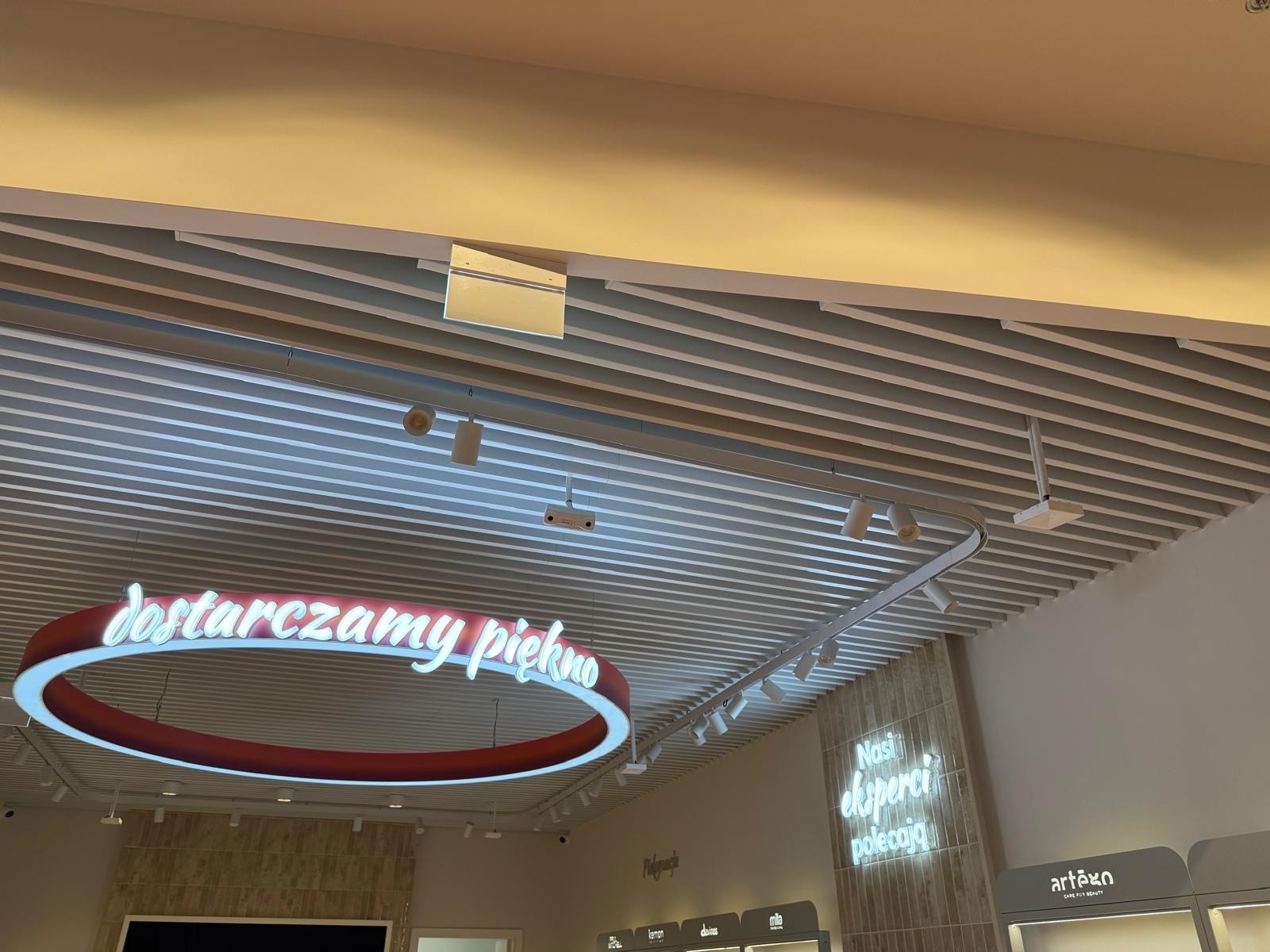 Sufit rastrowy z oświetleniem punktowym i neonowym napisem 'dostarczamy piękno'. Widoczne elementy instalacji i wykończenia wnętrza sklepu.
