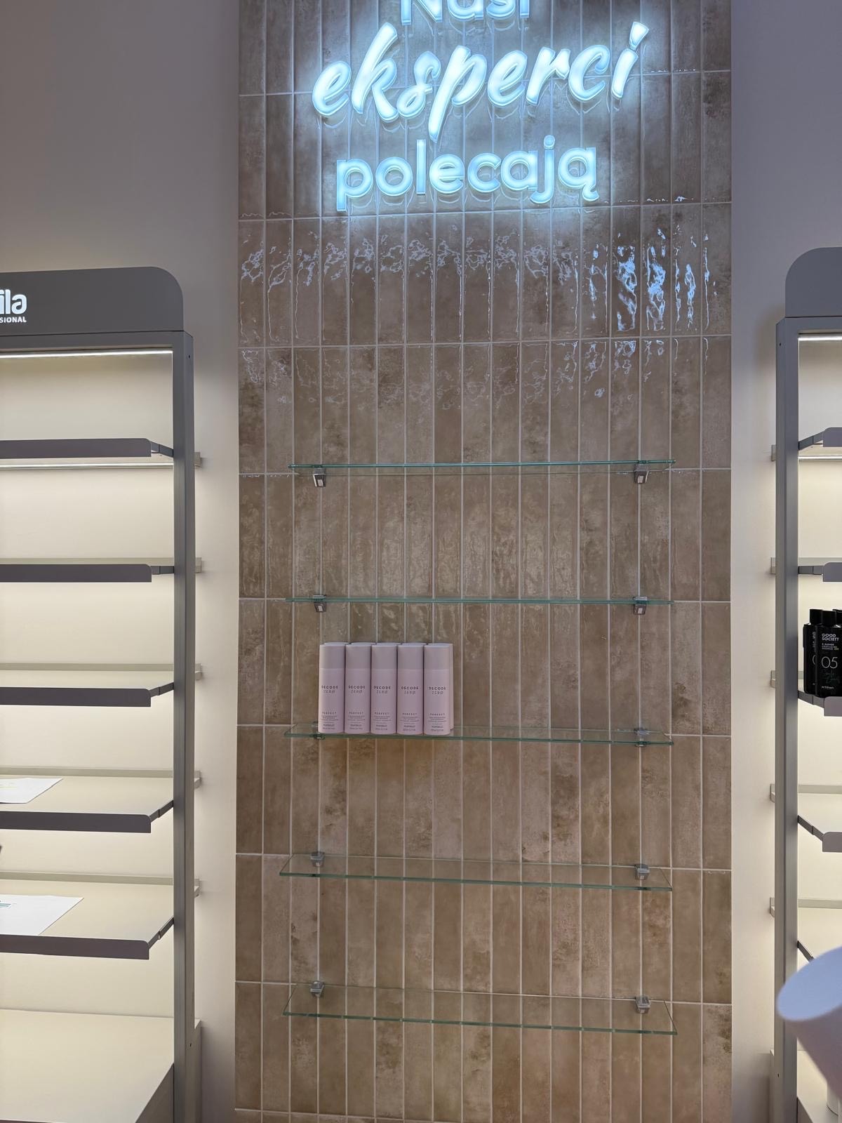 Beżowa ściana z płytek, podświetlony napis 'Nasi eksperci polecają', szklane półki z produktami. Widok na wykończone wnętrze salonu.