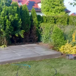 Wykonany taras z płyt w ogrodzie, otoczony zielenią i krzewami. Na trawniku widoczny robot koszący, a w tle dom z czerwoną dachówką.