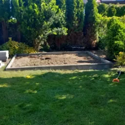 Przygotowany teren pod trawnik z rolki, obrzeża betonowe, narzędzia ogrodnicze na trawie. W tle iglaki i krzewy.