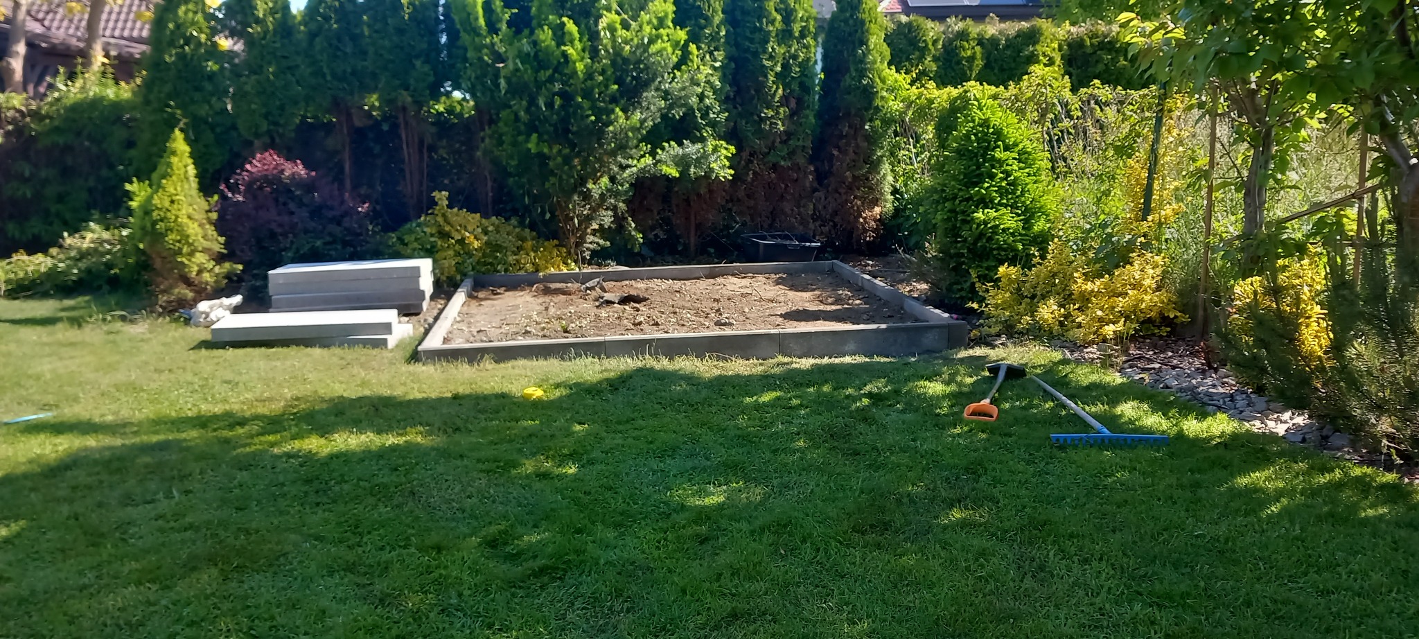 Przygotowany teren pod trawnik z rolki, obrzeża betonowe, narzędzia ogrodnicze na trawie. W tle iglaki i krzewy.