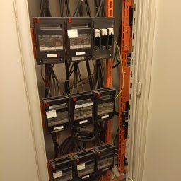 A-zabezpieczenia Adam Wysocki - Rozdzielnica elektryczna z przewodami i bezpiecznikami wewnątrz szafy. Widoczne oznaczenia na urządzeniach i kable w kolorach: czarnym, żółto-zielonym i brązowym.