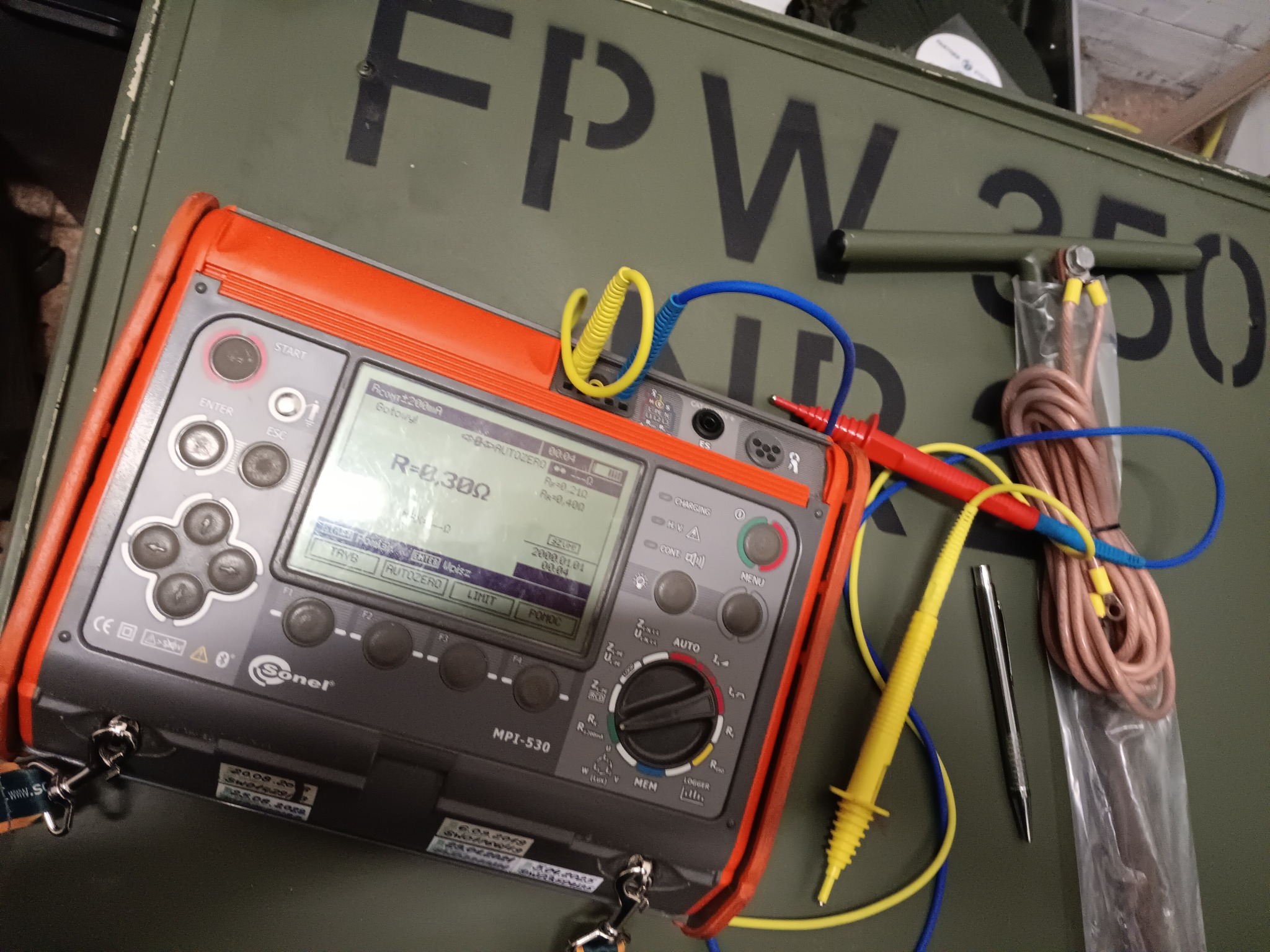 Miernik Sonel MPI-530 z przewodami pomiarowymi na tle skrzyni narzędziowej. Widoczne parametry pomiarowe na wyświetlaczu urządzenia.