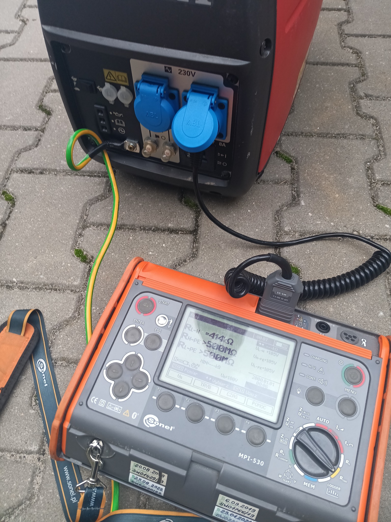 Generator prądu podłączony do miernika Sonel MPI-530 podczas testów. Widoczne kable, gniazda i wyświetlacz urządzenia pomiarowego z wynikami.