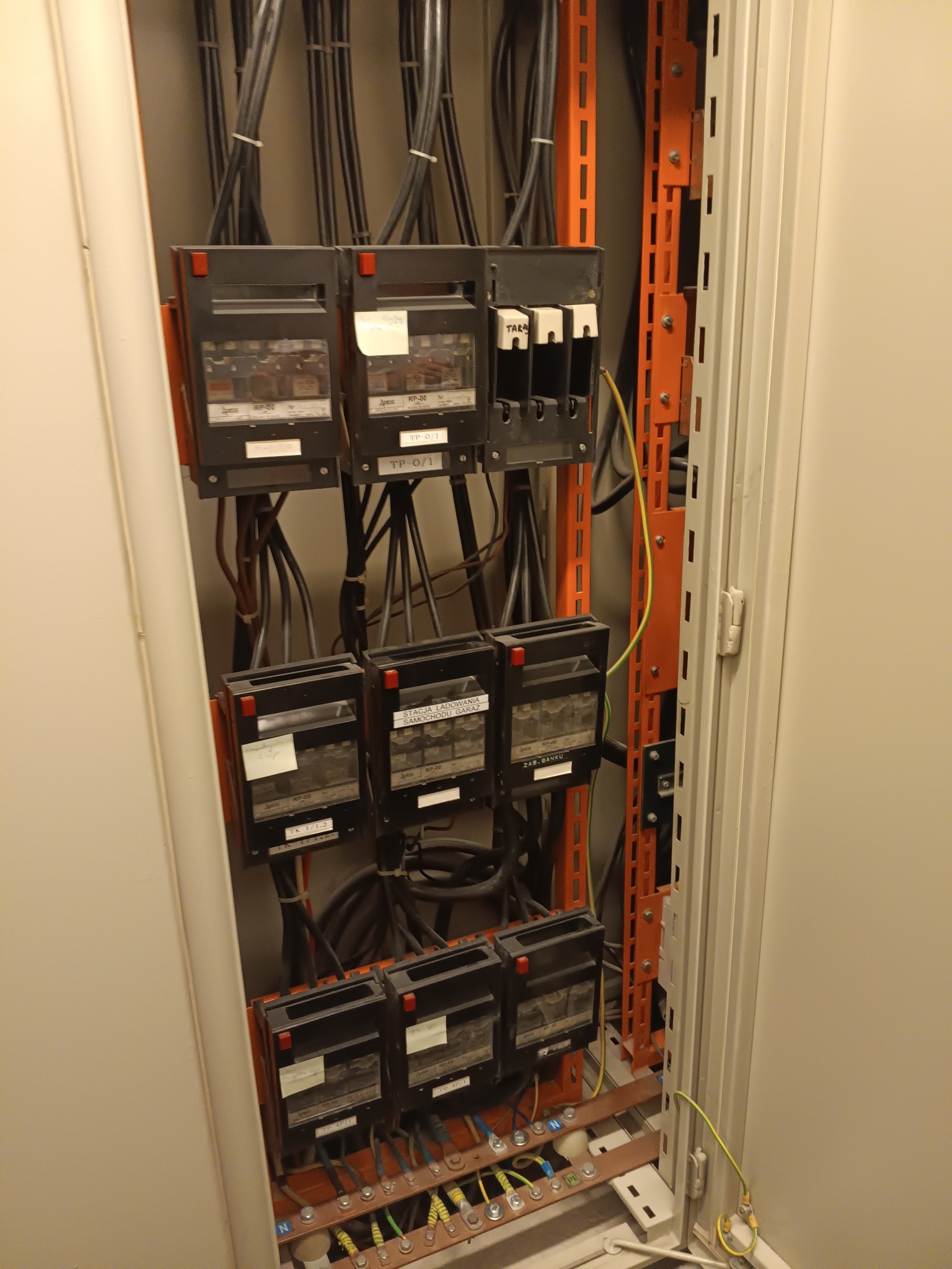 Rozdzielnica elektryczna z przewodami i bezpiecznikami wewnątrz szafy. Widoczne oznaczenia na urządzeniach i kable w kolorach: czarnym, żółto-zielonym i brązowym.