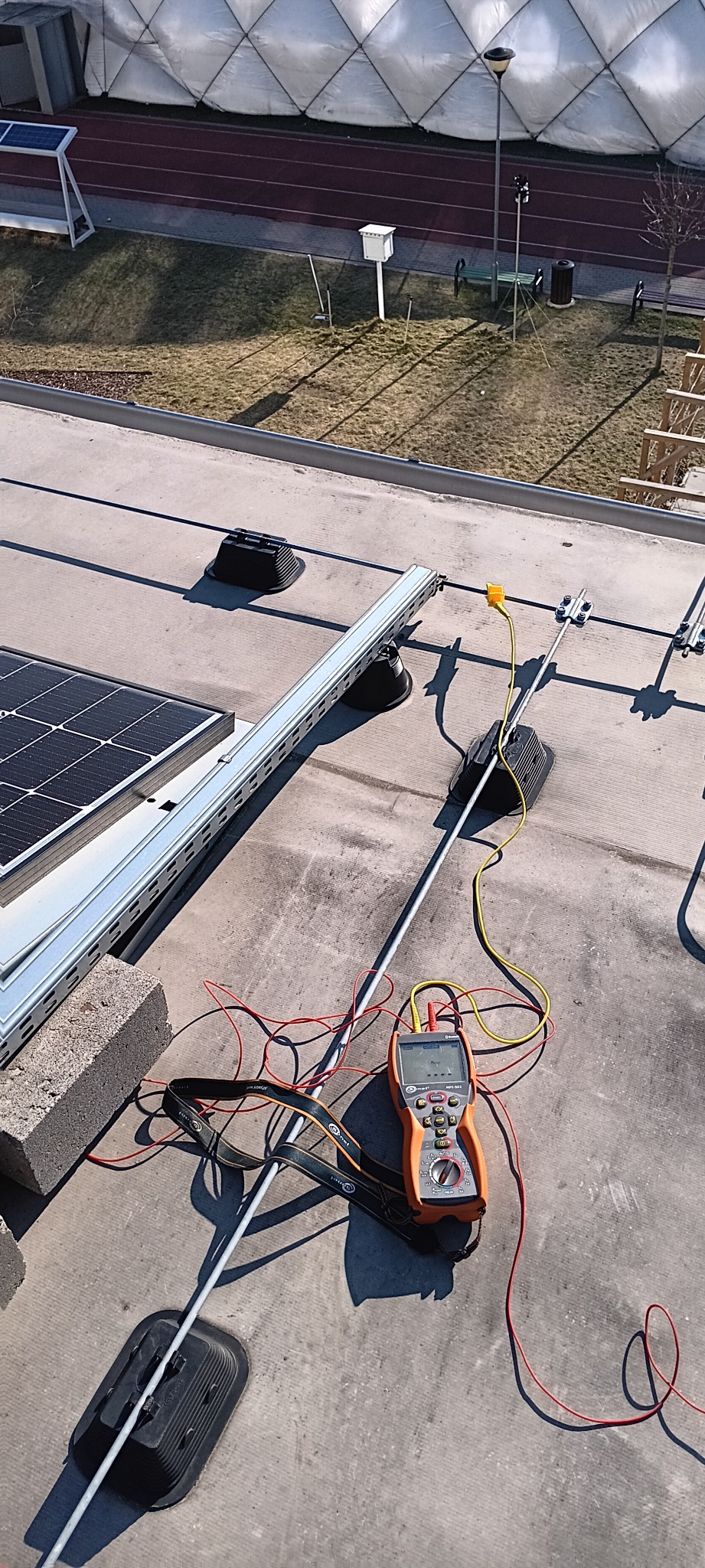 Pomiar instalacji fotowoltaicznej na dachu budynku. Widoczny miernik elektryczny z podłączonymi przewodami, elementy montażowe paneli i fragment panelu słonecznego.