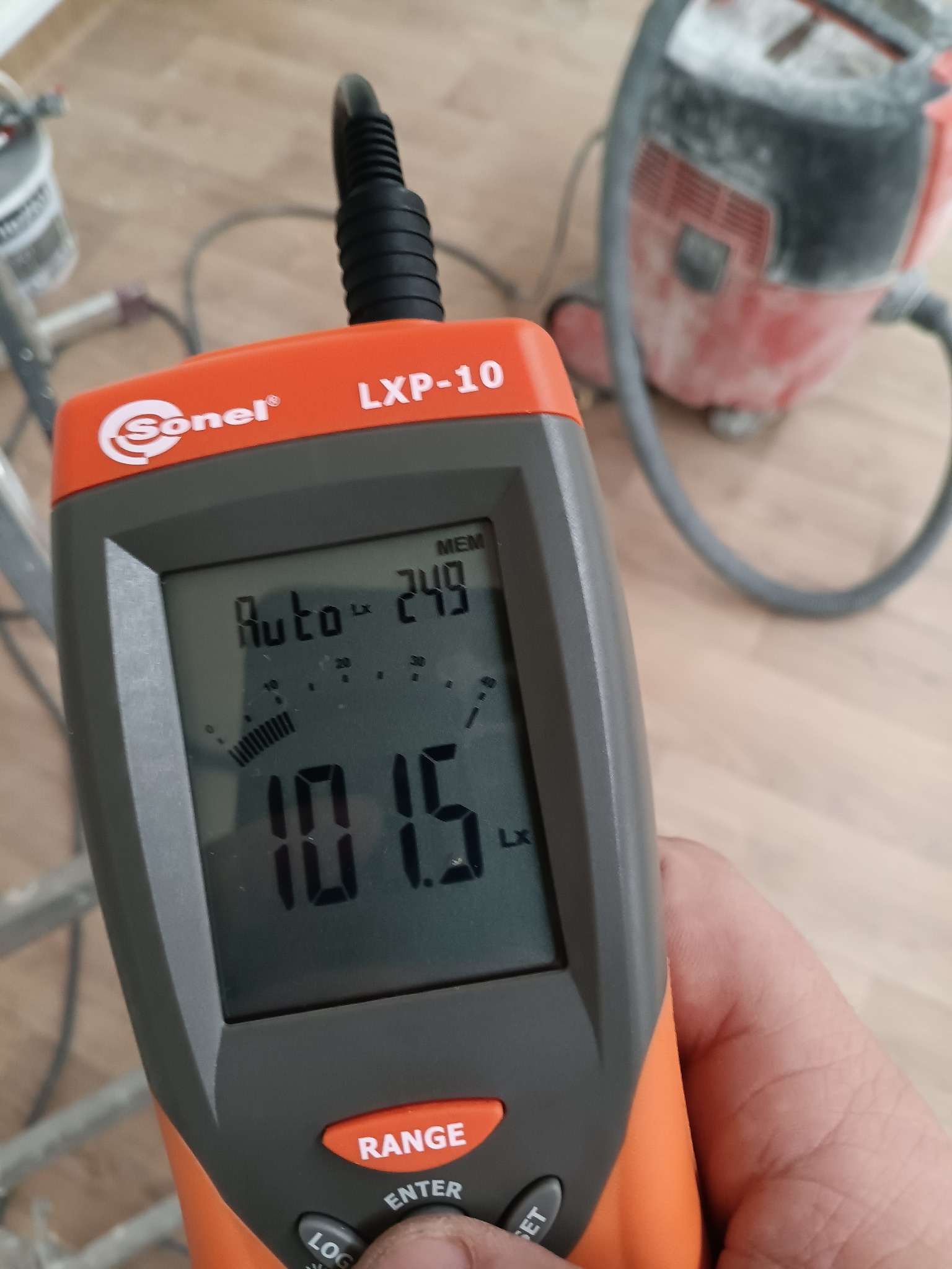 Miernik natężenia światła Sonel LXP-10 w dłoni, wskazujący 101.5 Lx, z odkurzaczem przemysłowym w tle. Pomiar oświetlenia w pomieszczeniu.