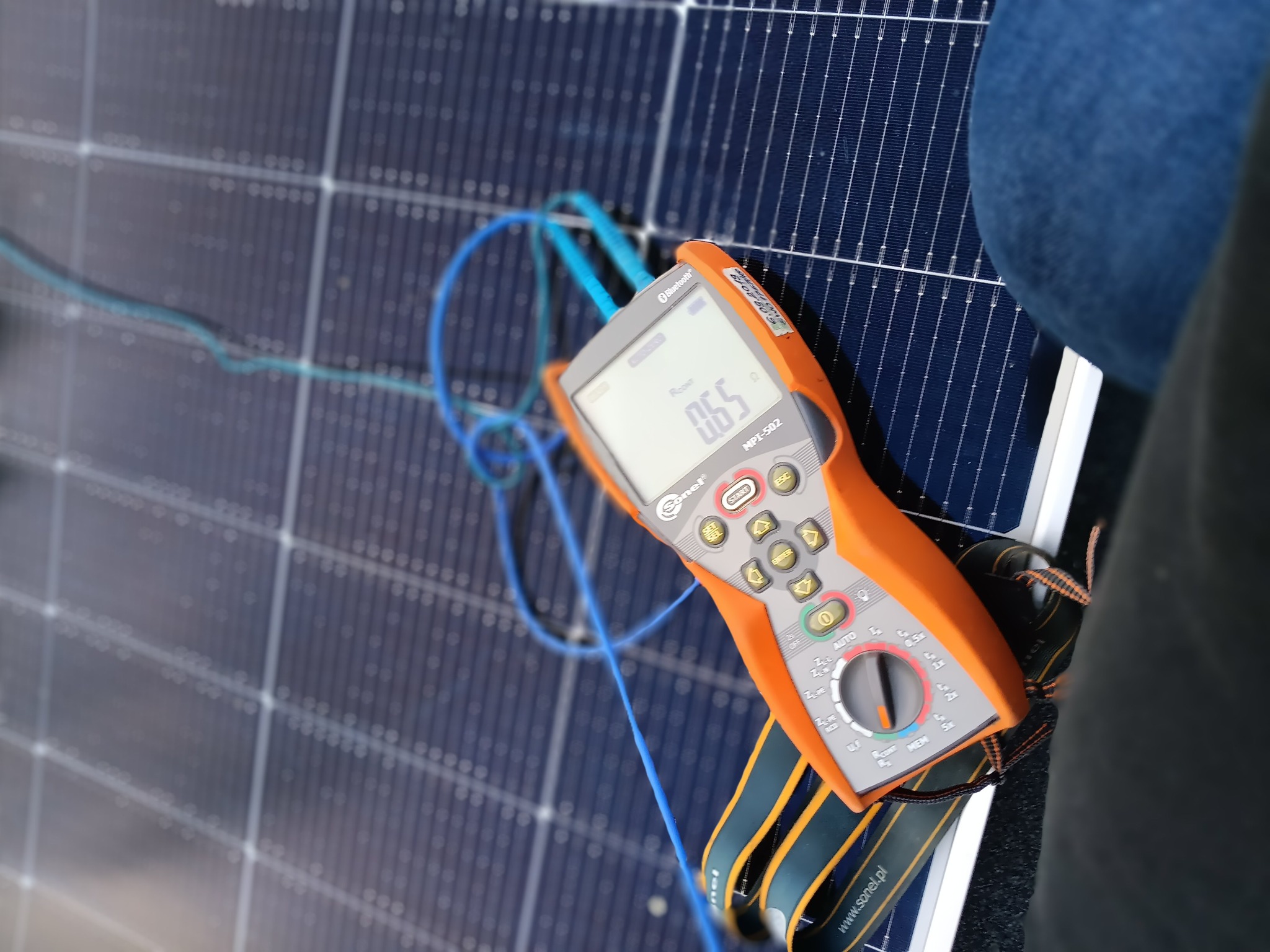 Pomarańczowy miernik Sonel MPI-502 z niebieskimi przewodami na tle niebieskiego panelu słonecznego. Widoczny fragment ubrania osoby wykonującej pomiar.