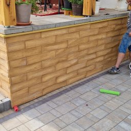 Adrian Tomczyk - Wykończenie elewacji imitacją drewna na cokole budynku z schodami. Widoczny pracownik z wiadrem farby i narzędziami. Detal wykończenia z bliska, ciepłe kolory.