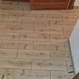 Ułożone panele podłogowe imitujące drewno w jasnym odcieniu, z widocznymi krzyżykami dystansowymi. Ujęcie z perspektywy wejścia do pomieszczenia.