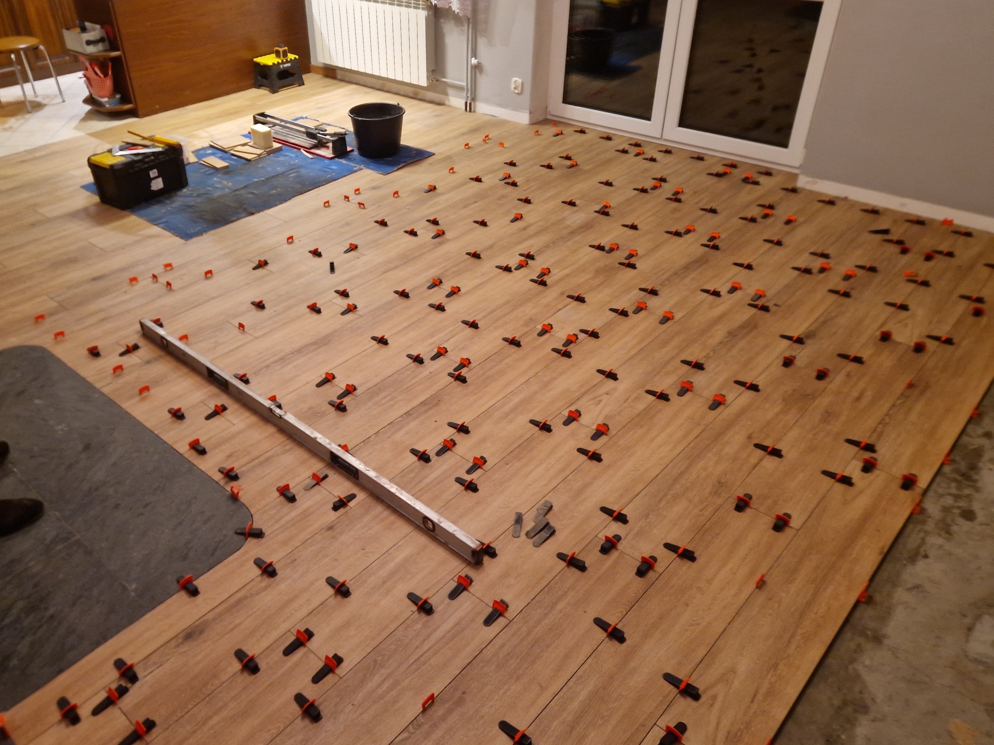 Układanie podłogi z paneli imitujących drewno. Widoczne narzędzia, poziomica i klipsy dystansowe. Częściowo ułożona podłoga z szarego materiału. Oświetlenie sufitowe.