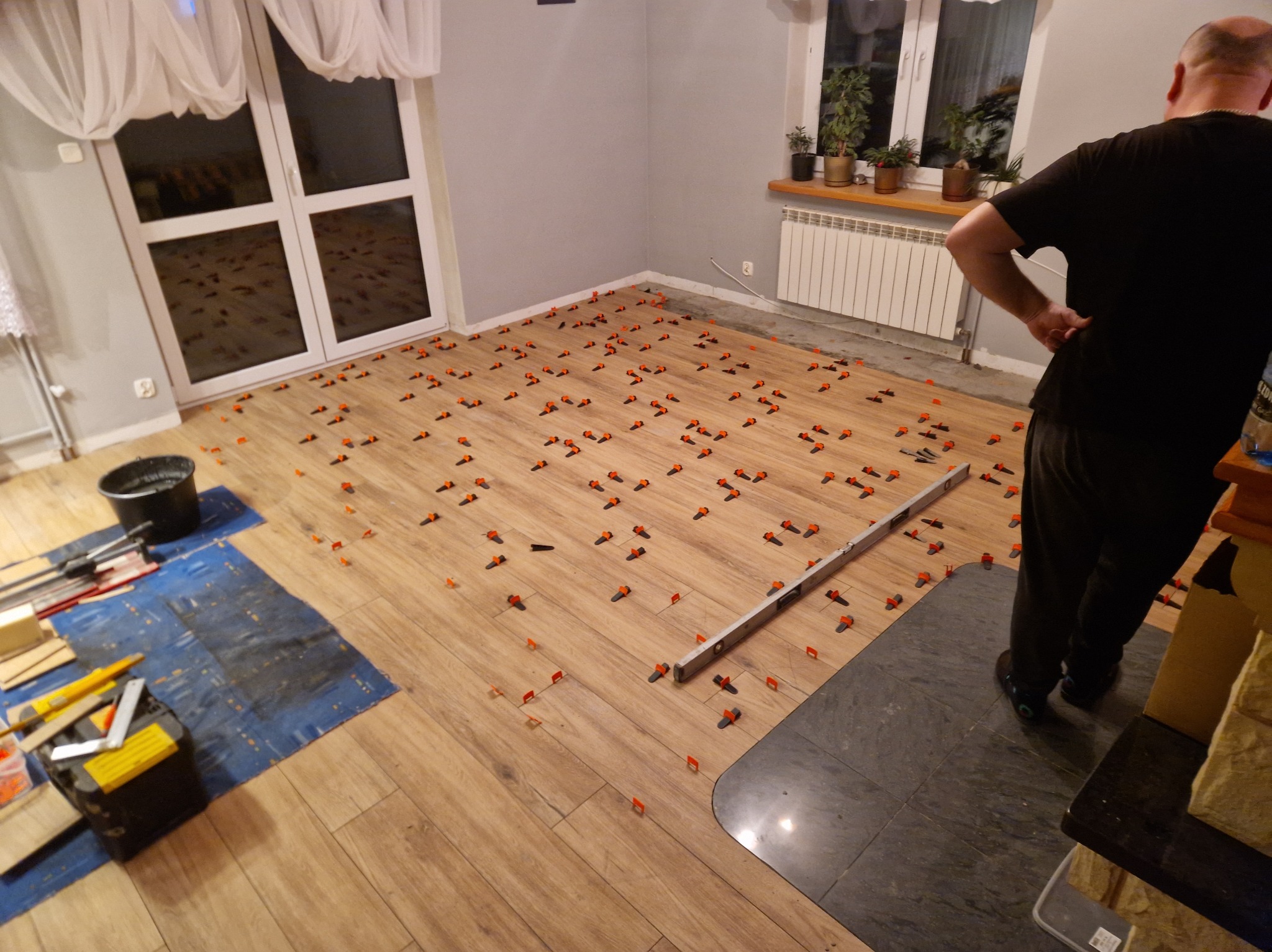 Proces montażu paneli podłogowych z użyciem klipsów poziomujących. Widoczne narzędzia i fragment ułożonej podłogi w pomieszczeniu z oknem i grzejnikiem.