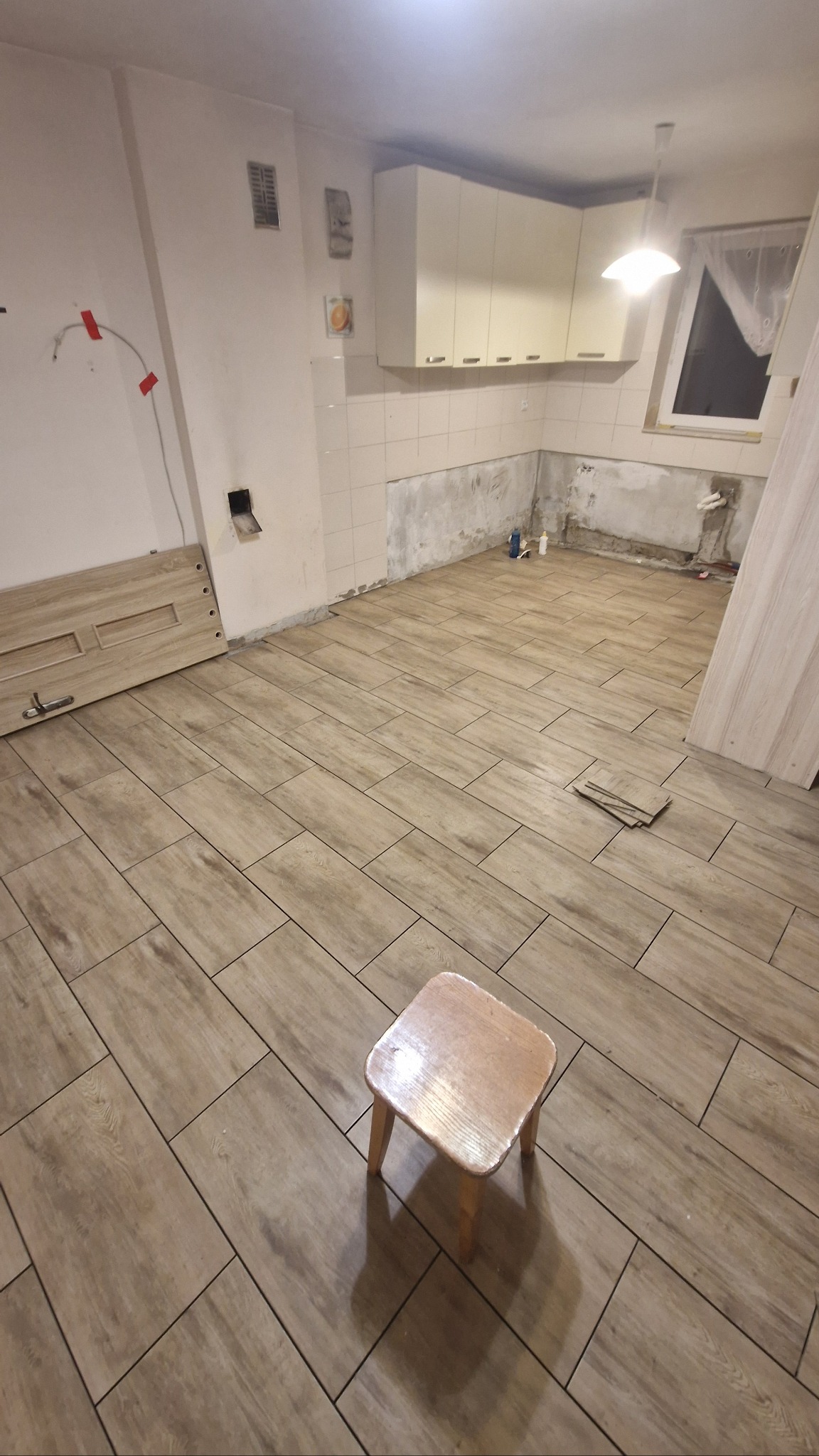 Układanie paneli imitujących drewno w kuchni, widoczny mały drewniany stołek. Częściowo położone płytki na ścianie. Praca w trakcie realizacji.
