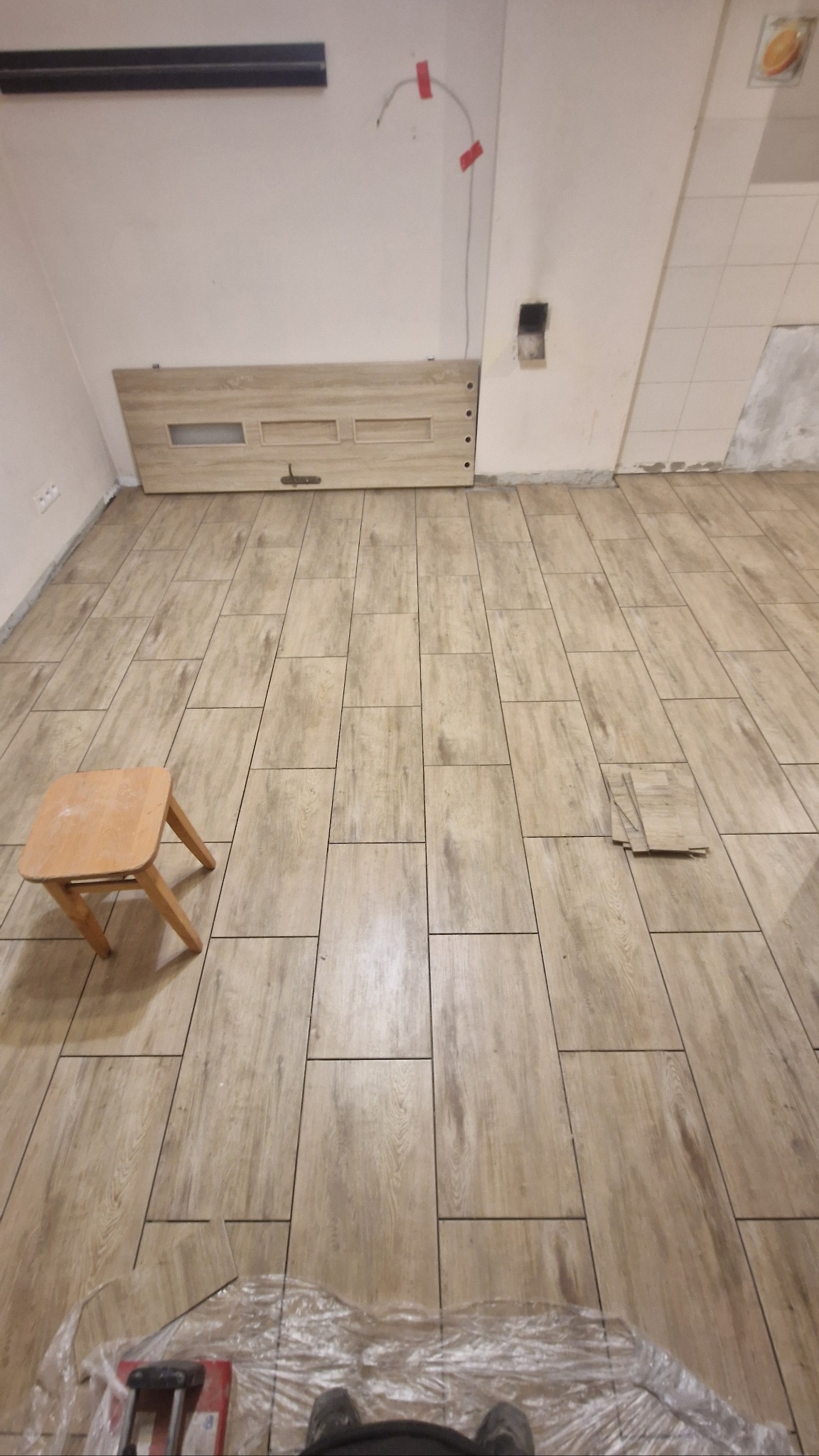 Jasne, prostokątne panele podłogowe imitujące drewno, ułożone w regularny wzór. W rogu mały stołek, przy ścianie leżące panele. Etap prac wykończeniowych.