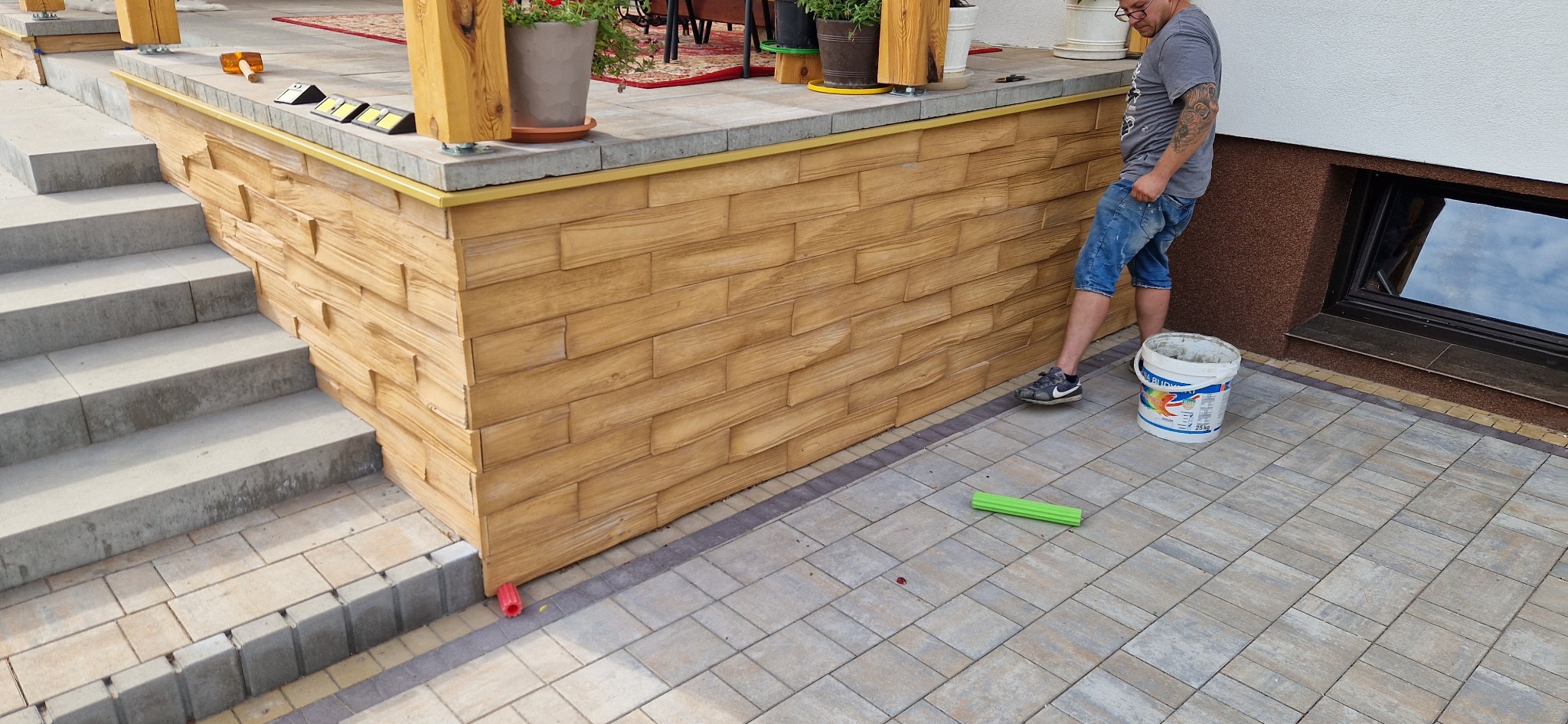 Wykończenie elewacji imitacją drewna na cokole budynku z schodami. Widoczny pracownik z wiadrem farby i narzędziami. Detal wykończenia z bliska, ciepłe kolory.