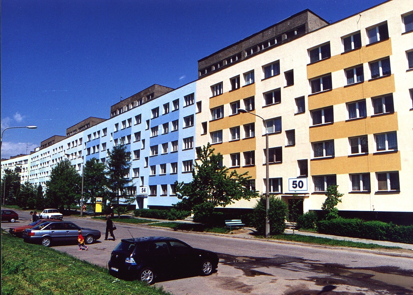 Wielobarwne bloki mieszkalne z numerami 50 i 54, auta na ulicy, drzewa i chodnik. Błękitne niebo.