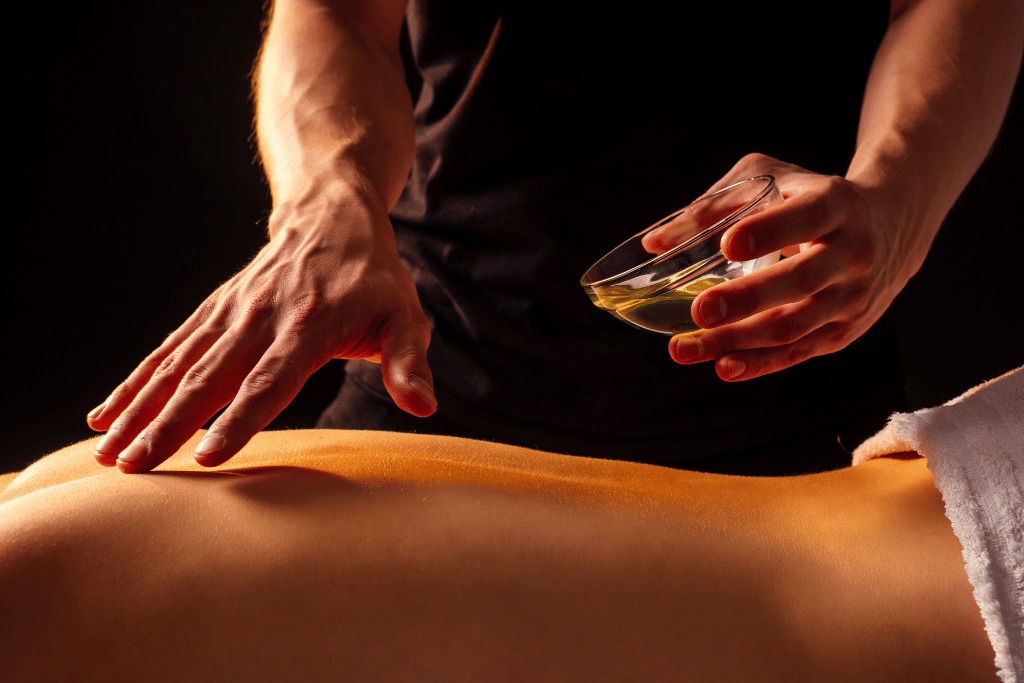 Olejkowy masaż pleców: dłoń masażysty aplikuje olej z miseczki na skórę klienta leżącego na stole z ręcznikiem, w ciepłym oświetleniu i ciemnym tle.
