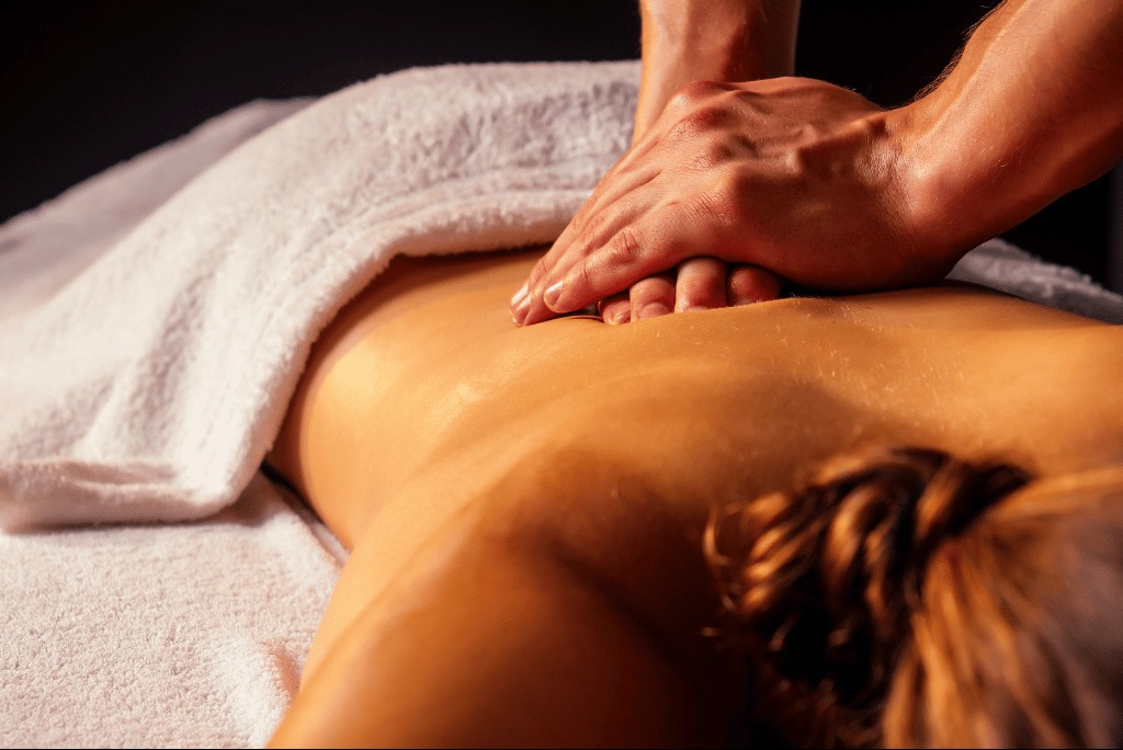 Dłonie masujące plecy kobiety leżącej na białym ręczniku, w ciepłym oświetleniu. Widoczne kręcone włosy. Skupienie na technice manualnej.