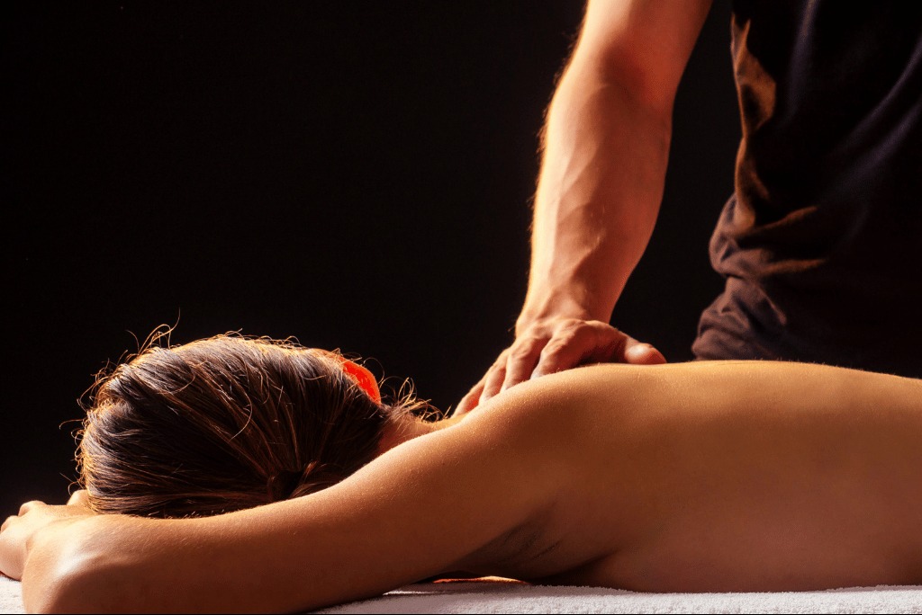Kobieta leży na brzuchu podczas sesji relaksacyjnej. Dłoń masażysty na plecach. Delikatne oświetlenie podkreśla napięcie mięśni. Ciemne tło.