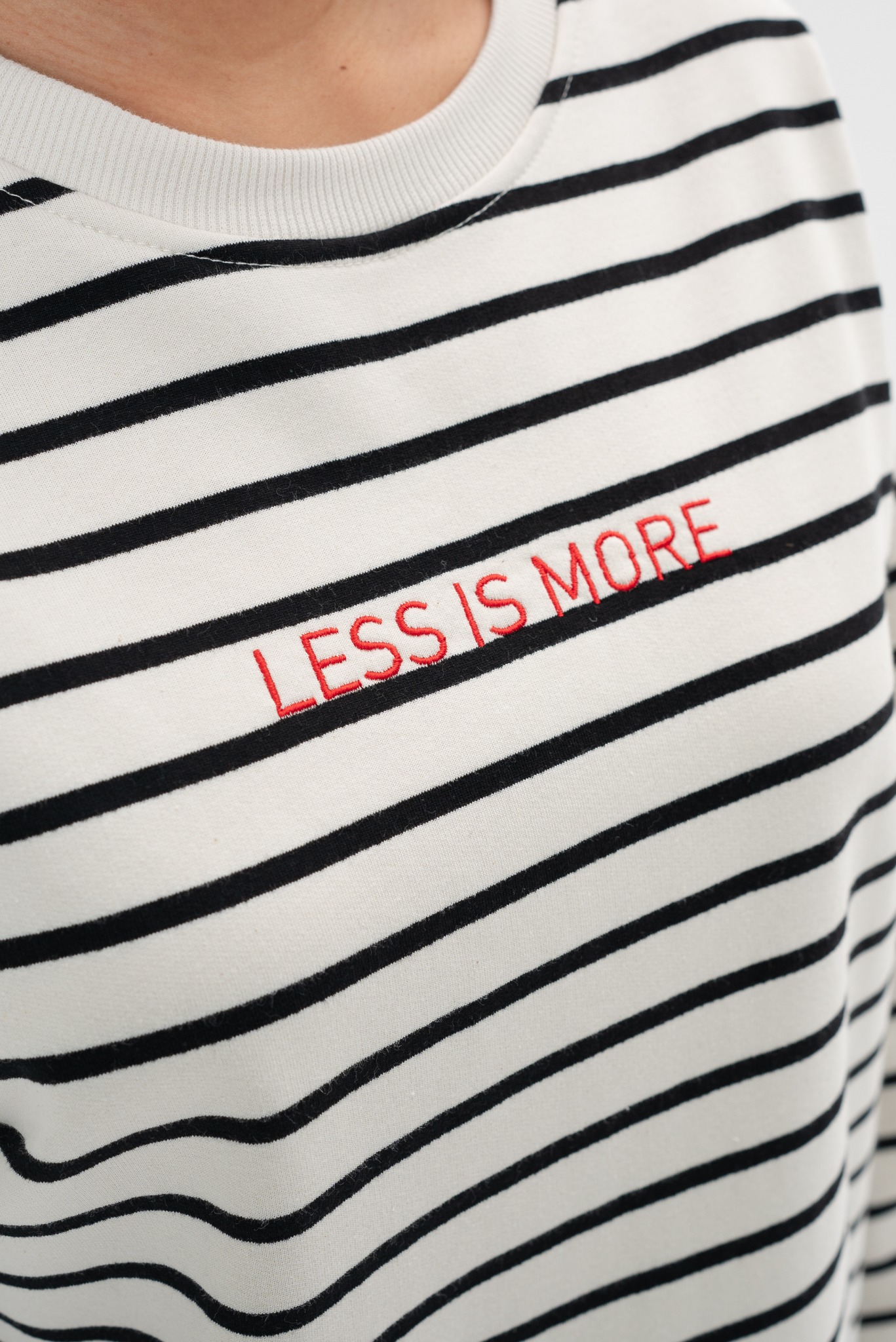 Paski na koszulce z czerwonym haftem 'LESS IS MORE'. Detal odzieży z bliska, prezentujący precyzyjny nadruk. Branding odzieży.