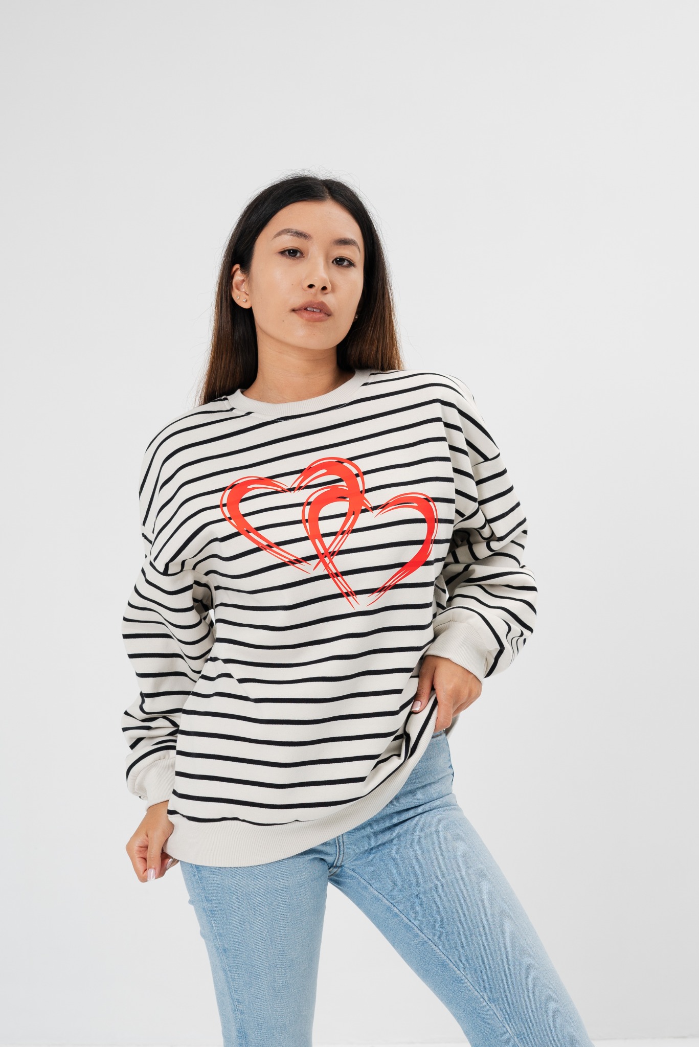 Modelka w bluzie w paski z czerwonymi sercami, prezentująca gotowy produkt. Jasne jeansy, białe tło. Wyraźny nadruk, naturalna ekspozycja.
