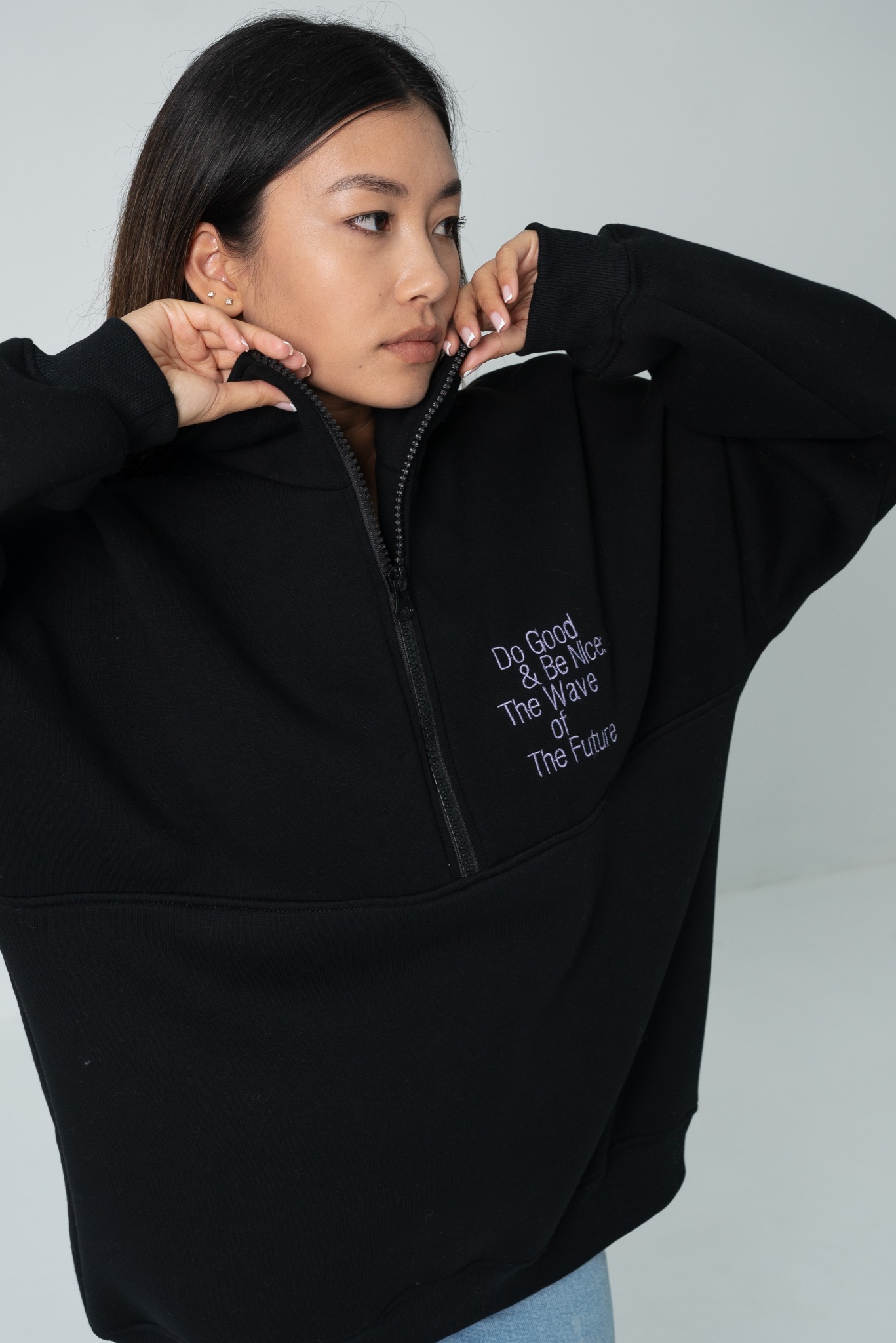 Kobieta w czarnej bluzie z napisem 'Do Good & Be Nice The Wave of The Future', zapinająca zamek. Widoczne delikatne kolczyki.