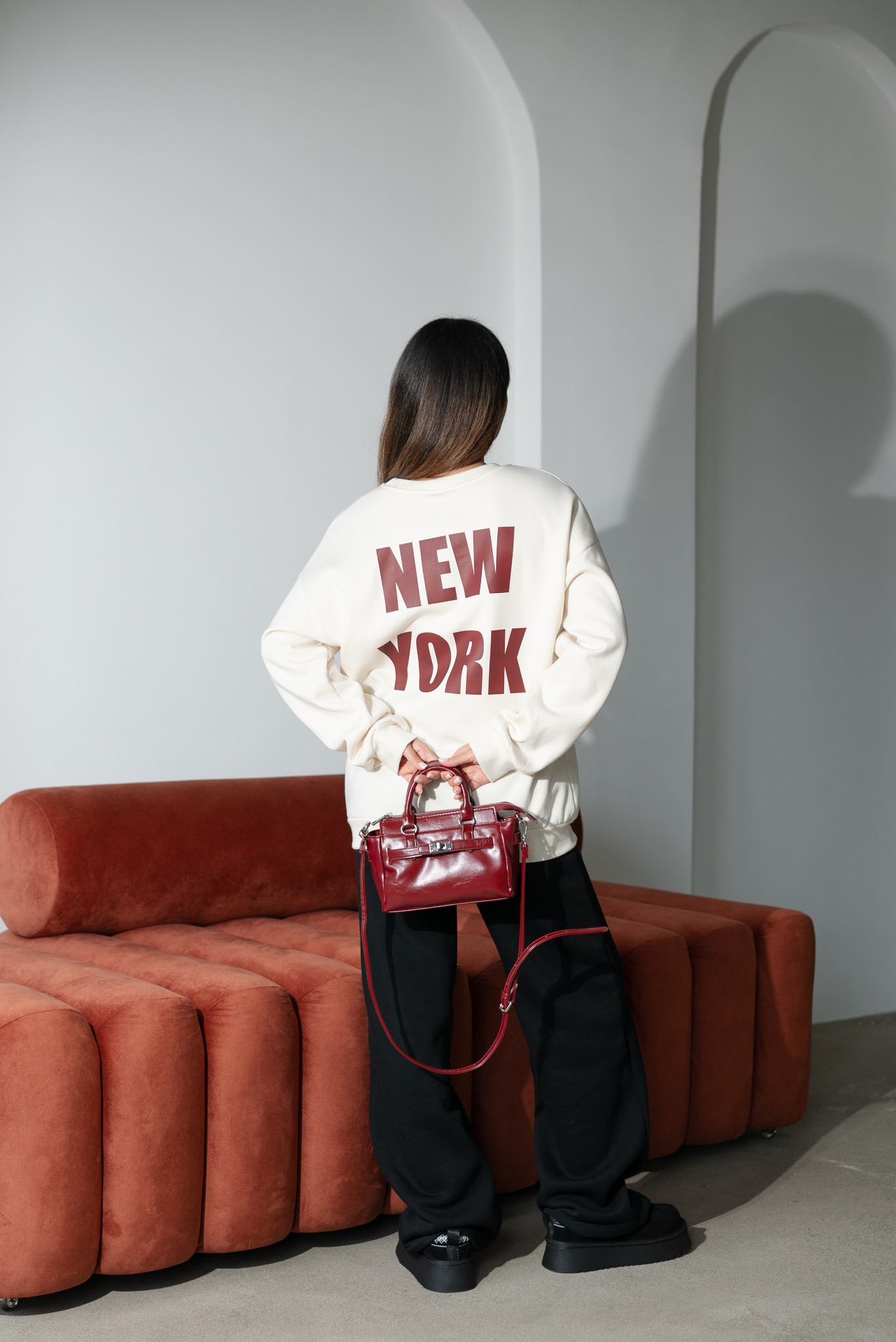 Osoba tyłem w bluzie z nadrukiem 'NEW YORK', trzymająca bordową torebkę na tle minimalistycznego wnętrza z kanapą.